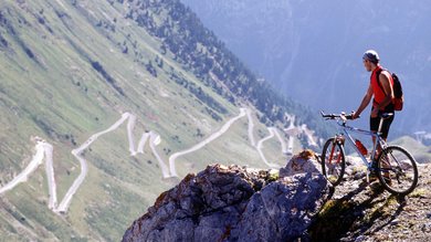 A mountain biker stands atop a peak, before him a breathtaking view of the serpentine passes of Vinschgau in South Tyrol.