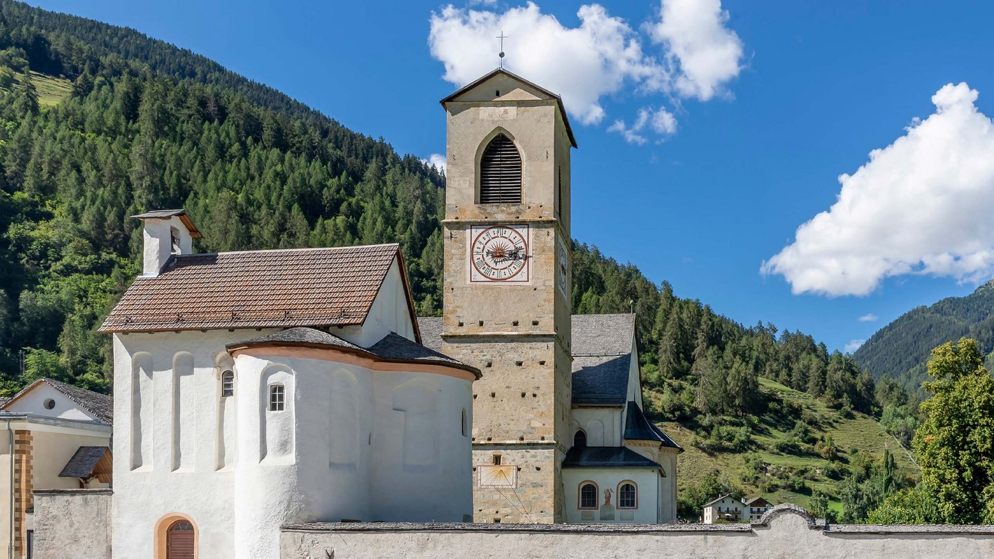 Die historische Kirche St. Johann in einem sonnigen Tal, mit ihrem charakteristischen Glockenturm, der sich gegen den blauen Himmel und die grünen Berghänge abhebt.