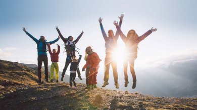 A group of friends joyfully leaps atop a mountain summit, framed by the warm glow of the sunset against the radiant blue sky.