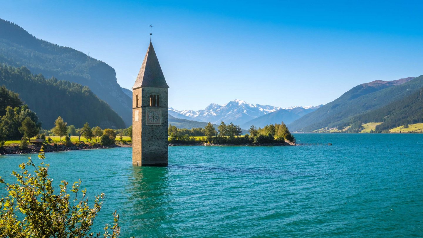 Der versunkene Kirchturm von Graun ragt aus dem klaren blauen Wasser des Reschensees, umgeben von leuchtend grüner Vegetation und der majestätischen Bergkulisse Südtirols.
