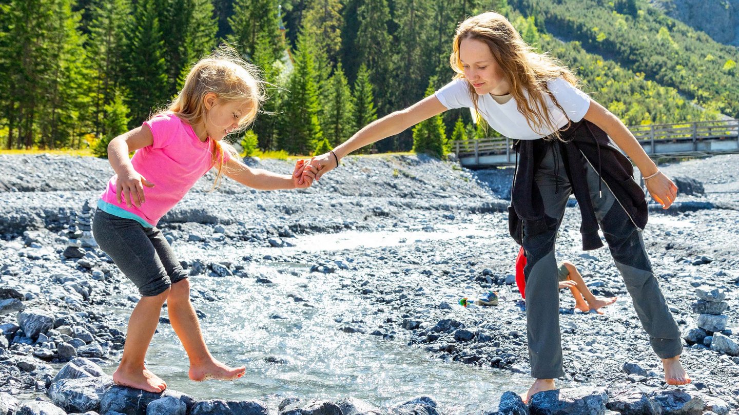 Two children play joyfully on a gravel-covered river bank in Vinschgau, helping each other balance across the sparkling stones.